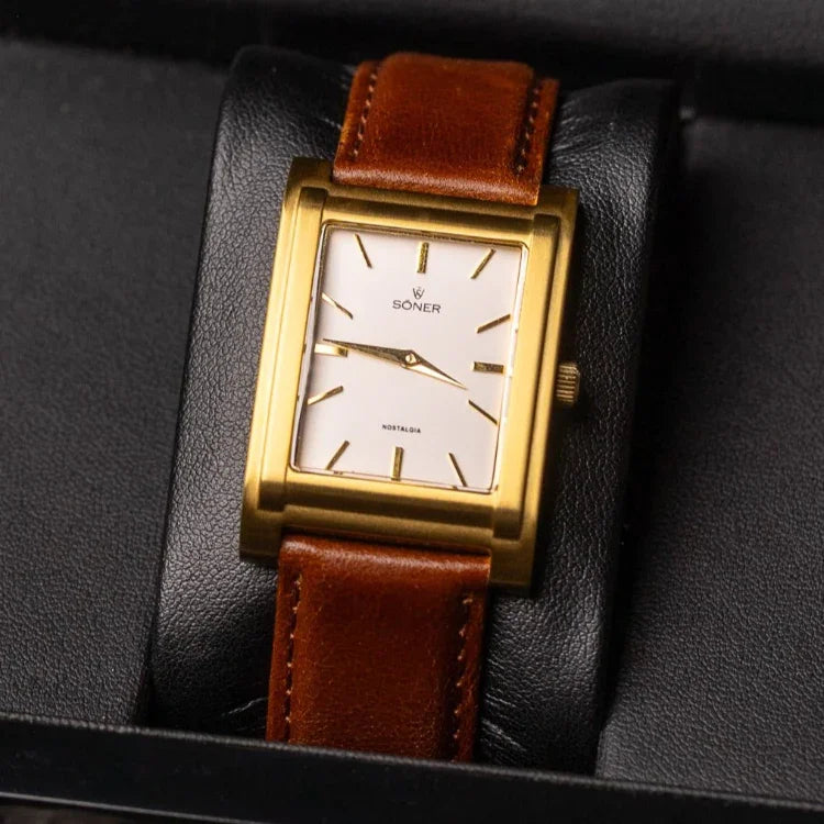 A rectangular watch gleams inside its black box, featuring a gold wristwatch with a brown leather strap. Its white face is adorned with simple black hour markers. The open box reveals a textured background underneath, enhancing the watchs elegance.