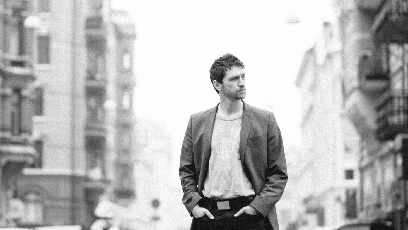 A man stands confidently in a city street, hands in his pockets. He wears a blazer over a t-shirt and sports a sleek rectangular watch as he looks to the side. The background is blurred, featuring buildings and cityscape details in monochrome.