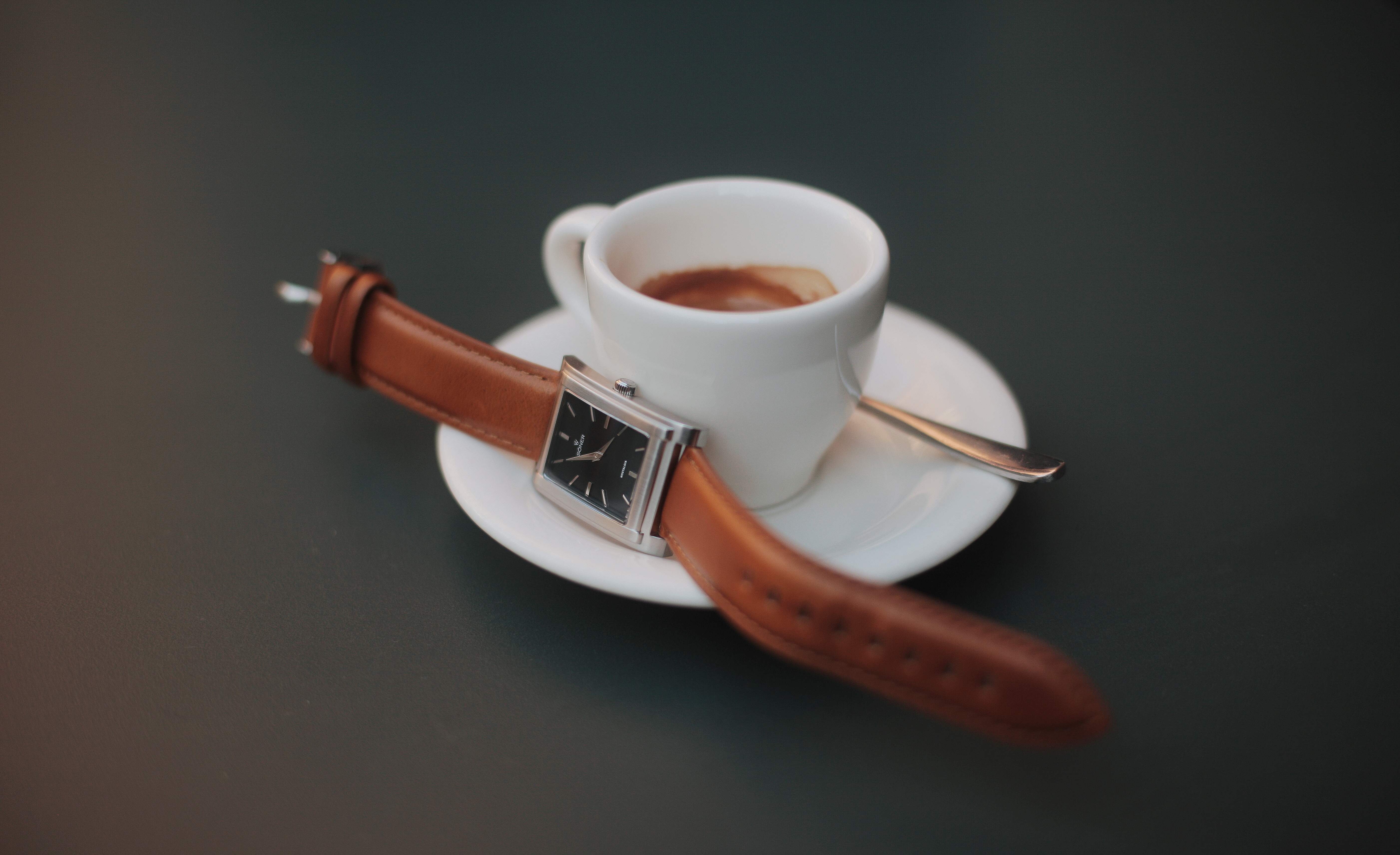 A white cup of espresso sits on a saucer with a spoon on a dark surface. A rectangular watch with a brown leather band is artistically wrapped around the cup, its face visible. The image exudes a warm, elegant feel.