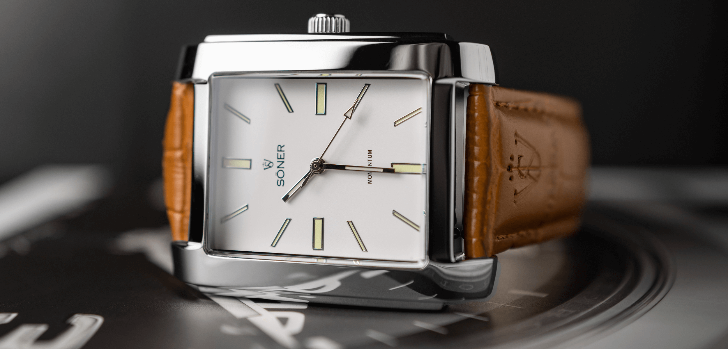 Close-up of a luxury rectangular wristwatch with a white face and silver accents. It features minimalist hour markers and the brand Soner on the dial. The watch has a brown leather strap and is set against a blurred dark background.