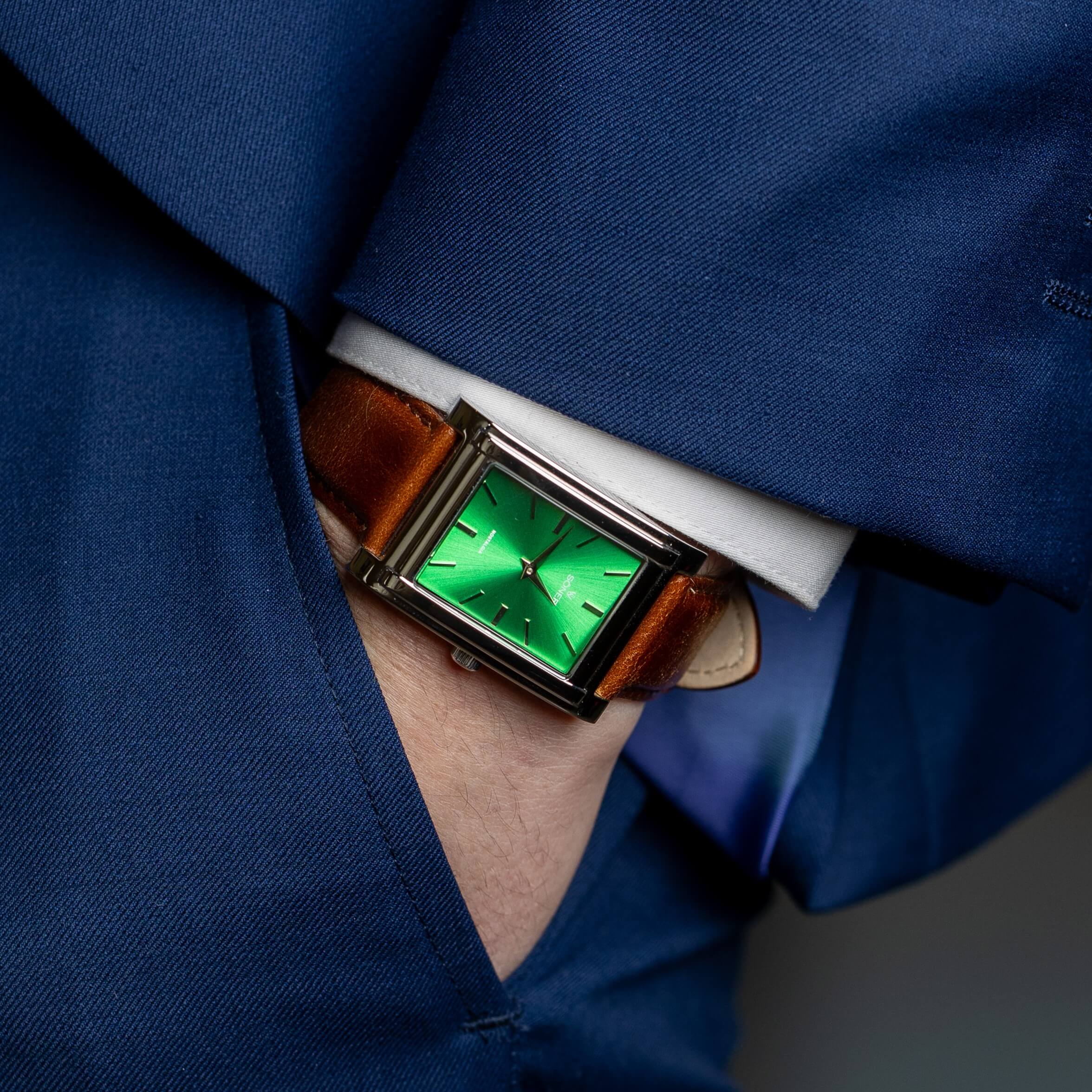 A person in a blue suit has their hand in their pocket, wearing a square, green-faced wristwatch with a brown leather strap. The watch has simple black hour markers and a sleek, modern design.