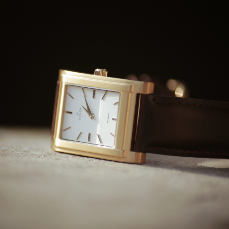 The rectangular gold wristwatch features a white face with minimalist black hour markers and hands, set against a soft-focus dark background. Its brown leather strap complements its elegant design, and the watch is artfully placed at an angle on a light surface.