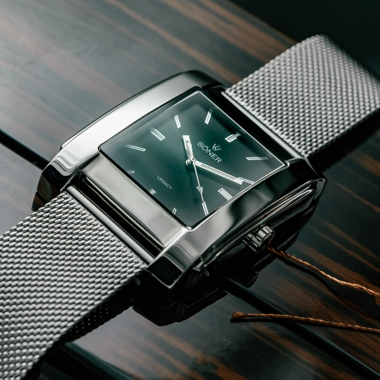 A sleek rectangular silver wristwatch with a black dial and mesh band sits elegantly on a glossy wooden surface. Featuring minimalist hour markers, its chic design highlights the brand SONER emblazoned on the face.
.