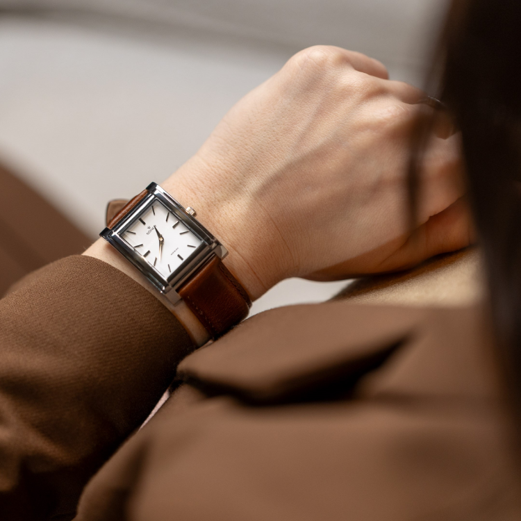 A person wearing a brown suit jacket is seen from the side, focusing on their wrist adorned with a rectangular watch featuring a brown leather strap. This timepiece boasts a simple, classic design with its white face and black hands.