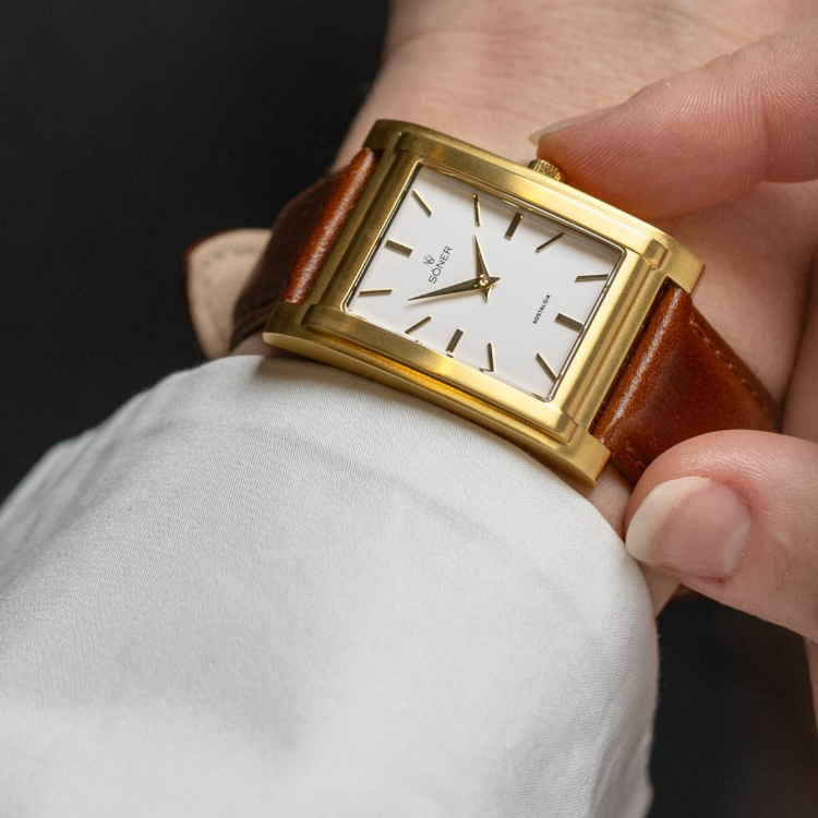 A close-up of a rectangular watch showcases its gold case and white dial on a brown leather strap. Thin black hour markers and two hands grace the face where the brand name is visible. A person in a white shirt is adjusting this elegant timepiece.