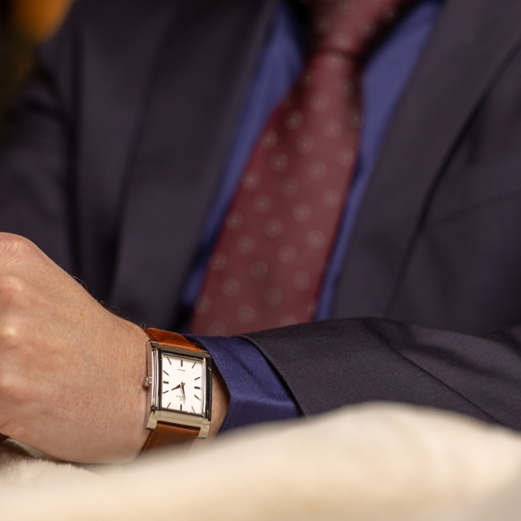 A person in a blue suit and burgundy polka dot tie is wearing a watch with a brown leather strap and a square, minimalist white face. The watch is in focus, showcasing its sleek design.