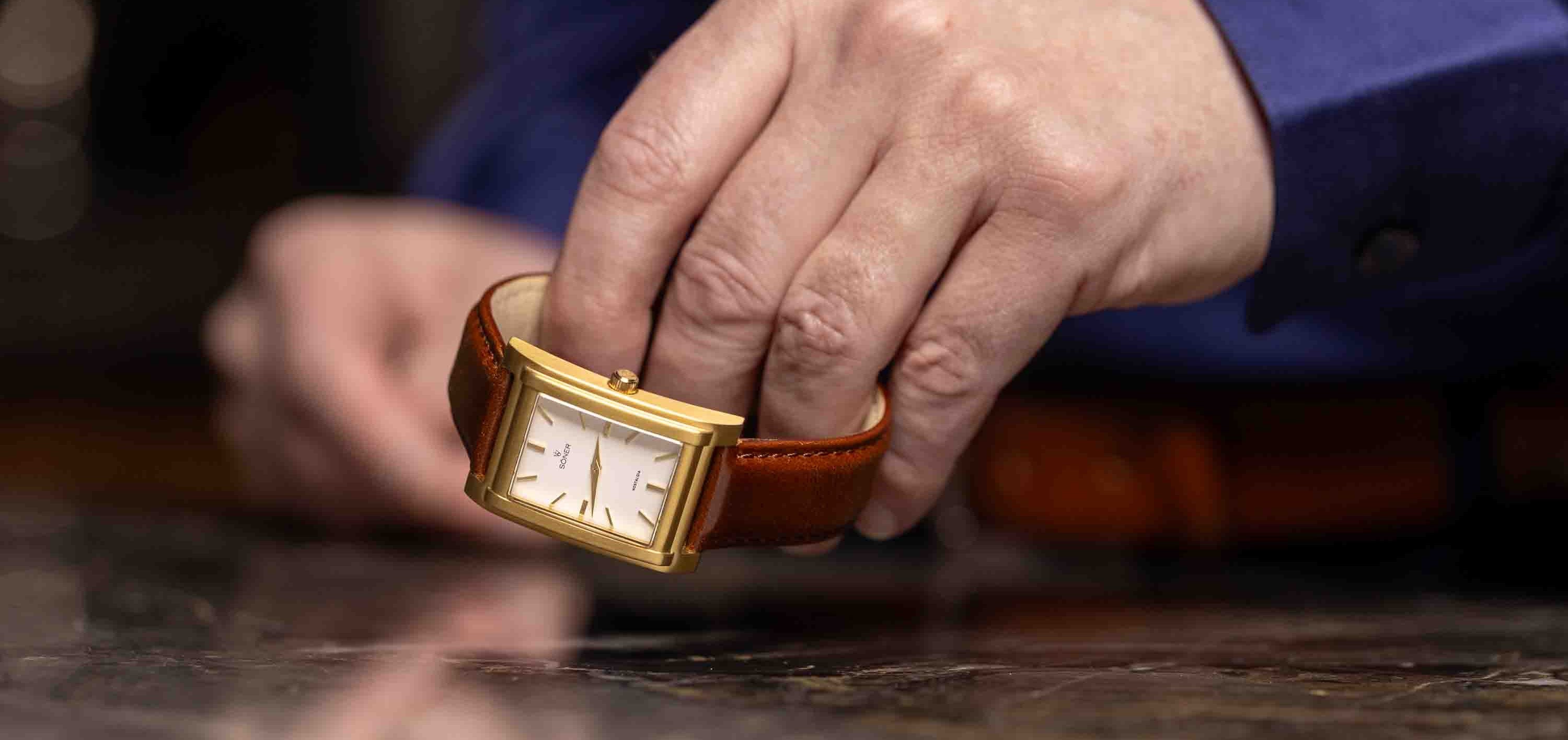 A person in a blue shirt is showcasing a striking rectangular watch, its gold frame gleaming. The timepiece rests over a reflective surface, highlighting its classic brown leather strap and elegant white face with minimalist markers.
