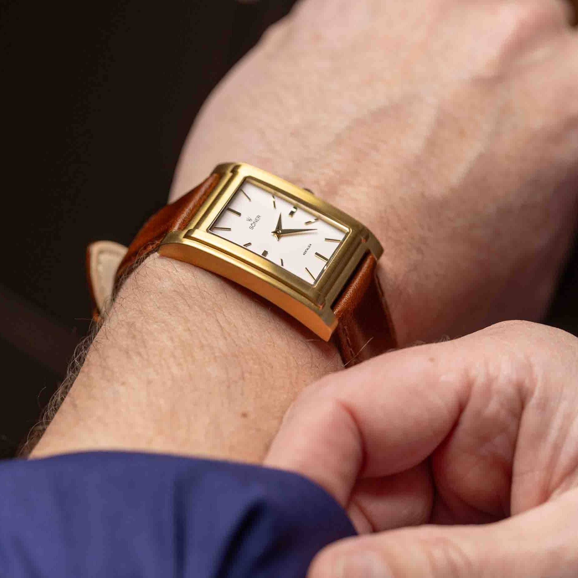 A person wearing the NOSTALGIA NICE with an 11-year battery checks the time. The gold rectangular watch features a white face, black hour markers, and a brown leather strap.