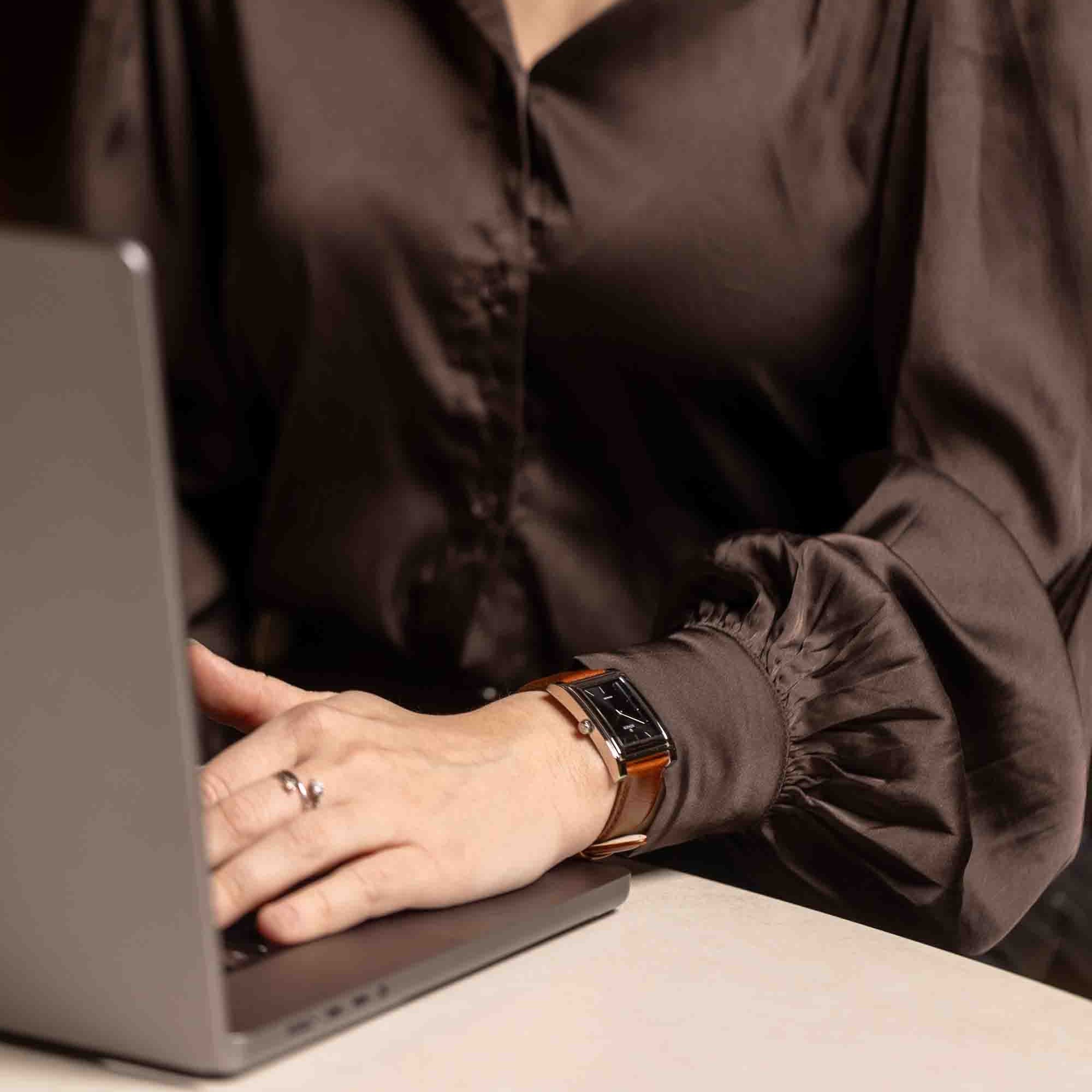 A person wearing a brown blouse types on a laptop. They are wearing a smart watch with a brown leather strap on their left wrist.