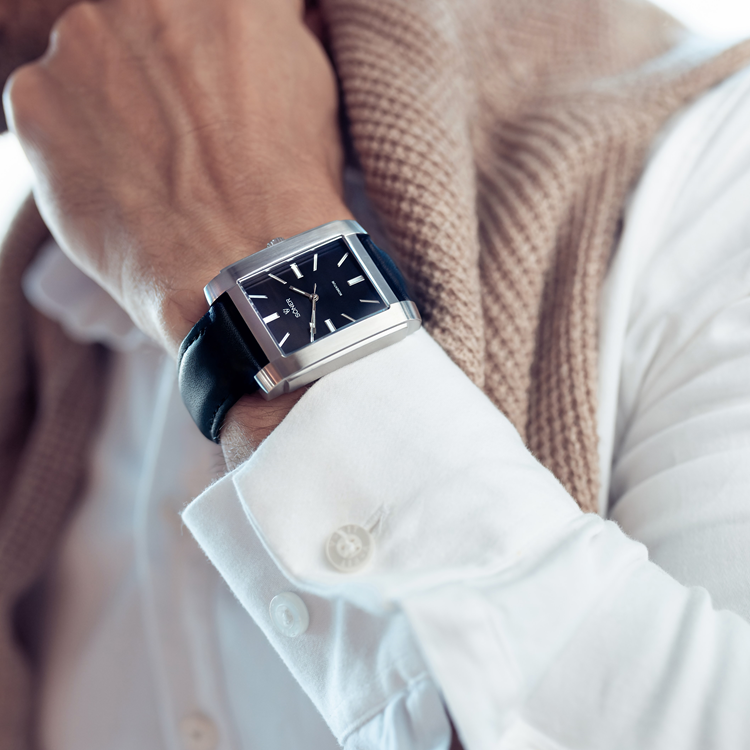 A person wearing a stylish rectangular watch with a black leather strap complements their white shirt and the beige sweater draped over their shoulders, casually resting a hand on their neck. The watch face is dark with silver markers.