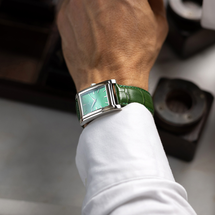 A person wearing a stylish rectangular watch with a green face and silver casing pairs it elegantly with a white shirt, their hand resting on a surface while a vintage camera sits in the background.