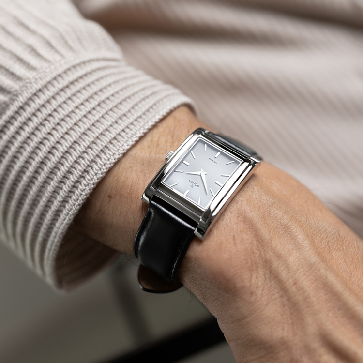 A person wearing a rectangular watch with a black leather strap and silver face, displaying the time. They are dressed in a ribbed, light-colored sweater. The minimalist design of the rectangular watch stands out against the soft texture of the sweater.