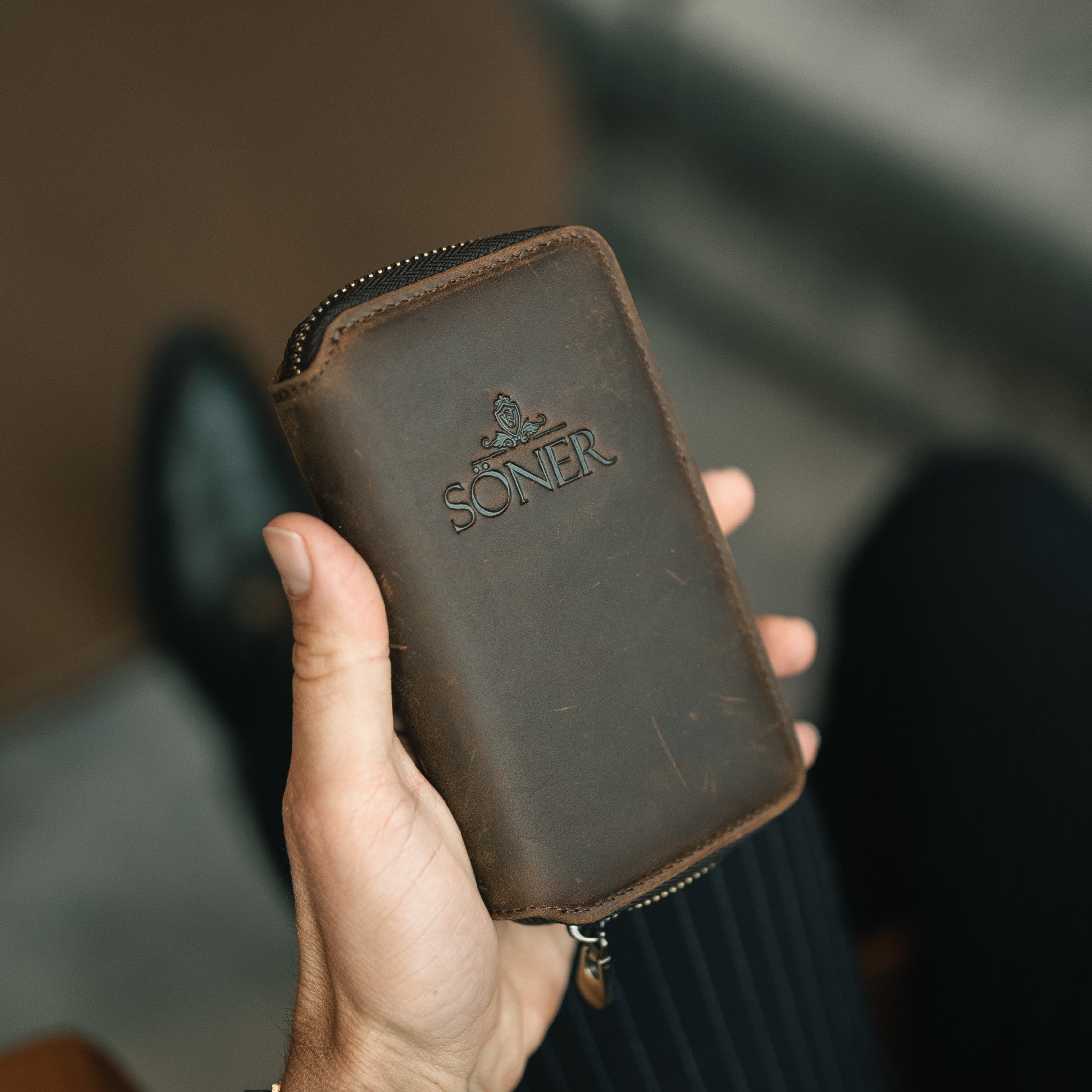 Hand holding a brown leather wallet with 'Soner' branding against a blurred background