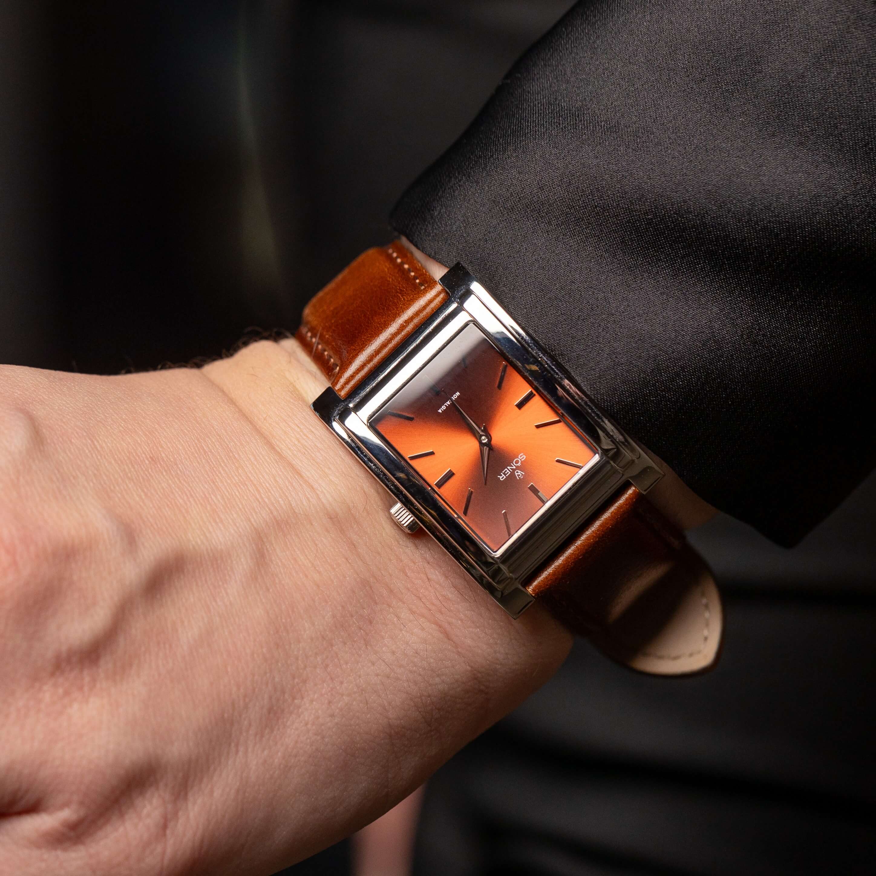 A rectangular wristwatch with a brown leather strap and a warm amber face, featuring minimalistic hour markers, is worn on a persons wrist, partially covered by a black suit sleeve.