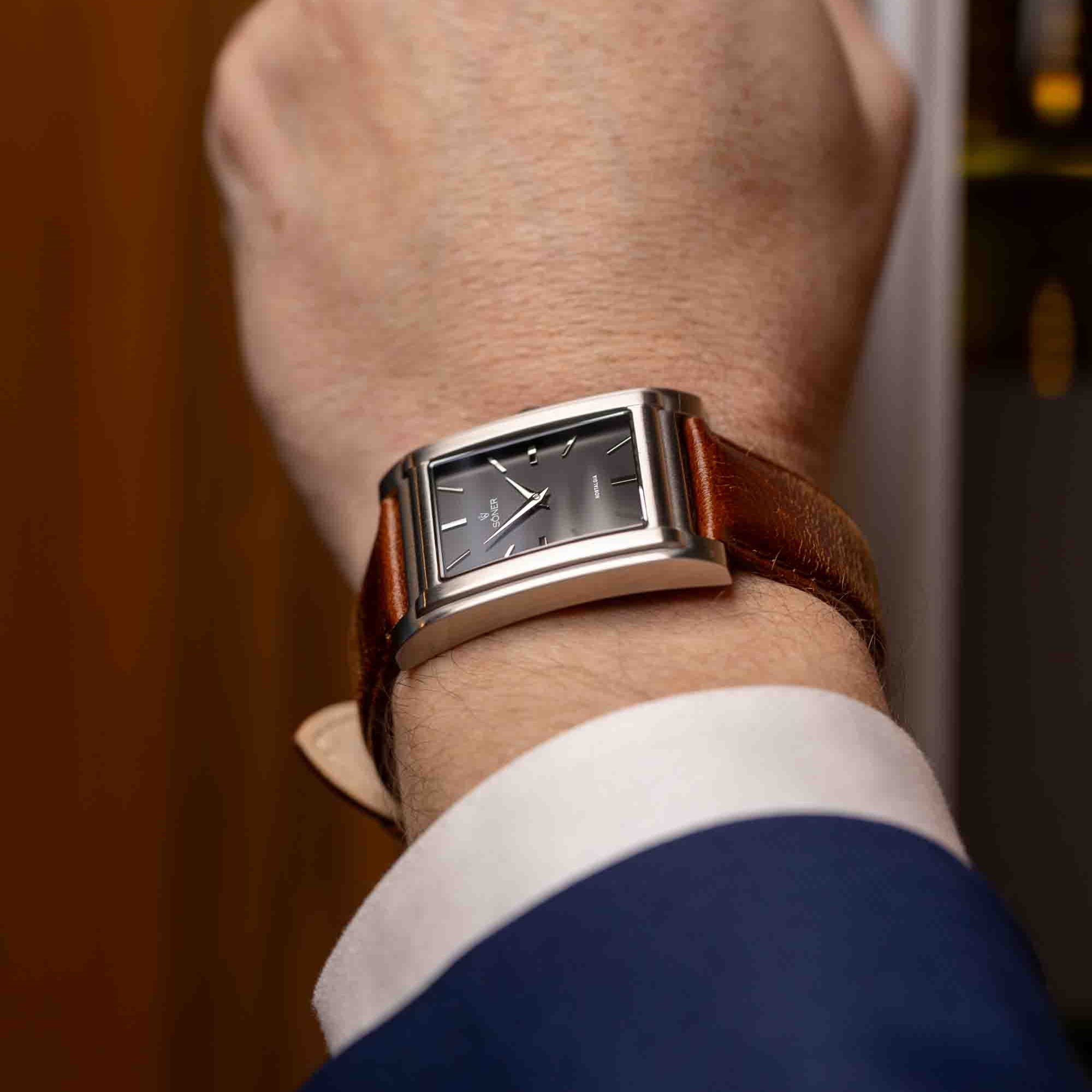 A person wearing a sleek rectangular watch with a silver case and brown leather strap on their wrist. The dark watch face, marked minimally, complements the blue suit and white shirt ensemble perfectly.