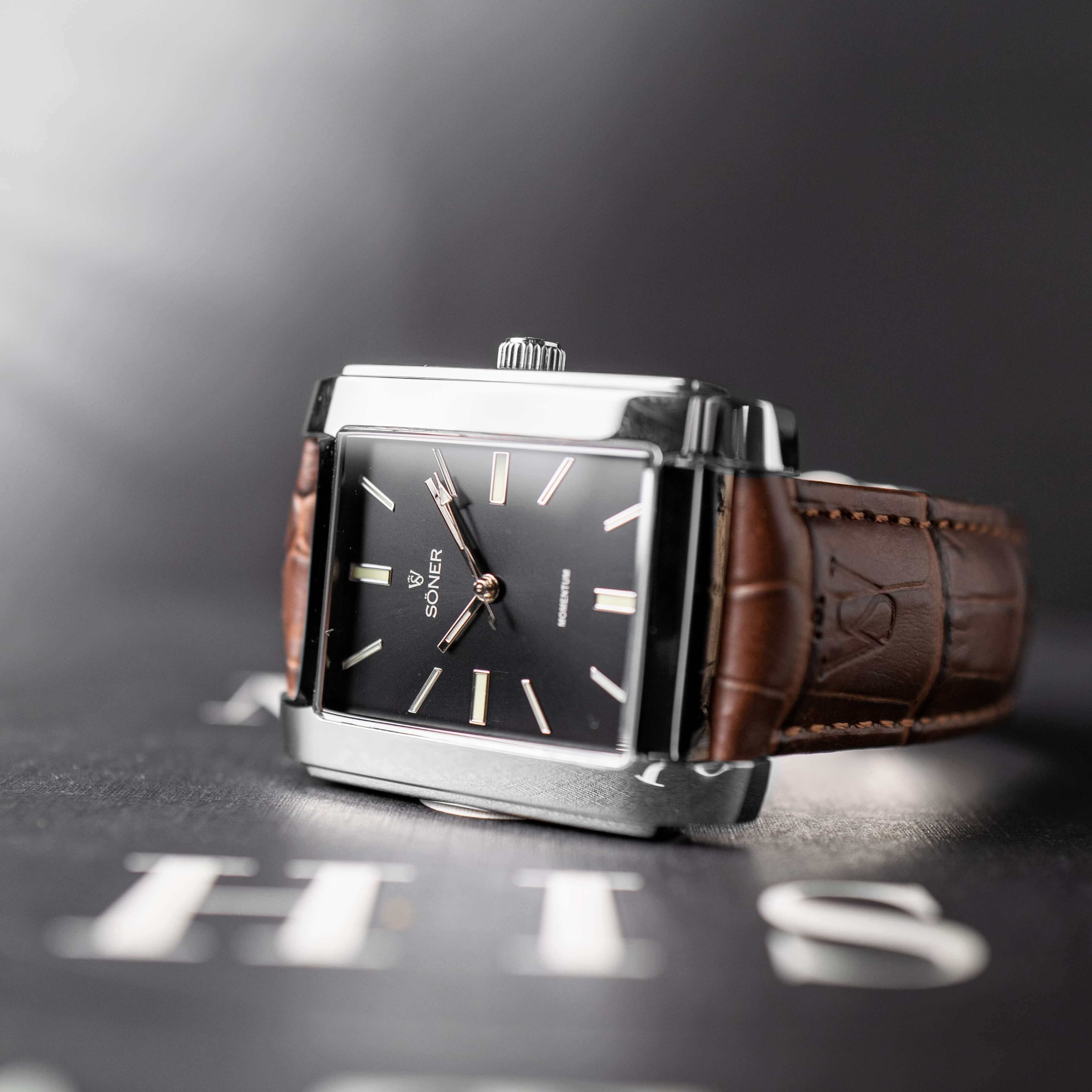 A close-up shot of a rectangular, silver-framed wristwatch with a black face and brown leather strap. The watch is angled on a dark surface with blurred text partially visible. The lighting creates subtle reflections on the metal.