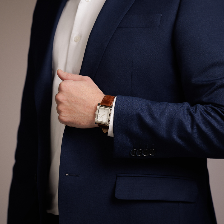 A person in a blue suit and white shirt adjusts their cuff, revealing a wristwatch with a brown leather band. The focus is on the suit details and the watch.