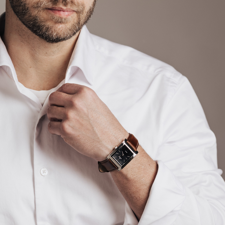 A person wearing a white button-up shirt adjusts their collar. They have a brown leather watch with a rectangular black face on their wrist. The background is a plain, neutral color.