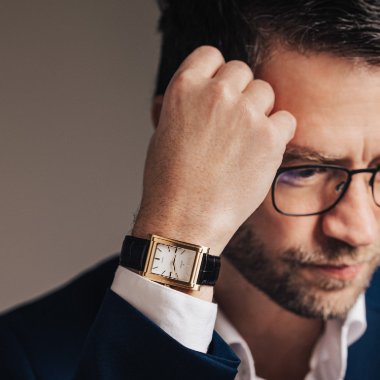 A man with glasses, dressed in a dark blue suit, is adjusting his rectangular watch with his right hand. The timepiece features a gold frame and black leather strap. The softly blurred background highlights both the watch and the mans intent expression.