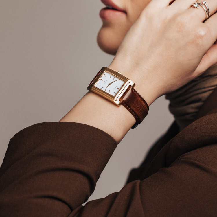 Close-up of a person wearing a rectangular gold wristwatch with a brown leather strap. The watch face is white with black hands and markers. The person wears a brown jacket and a scarf, with their hand touching their neck.