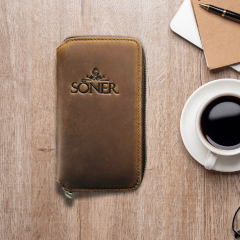 A brown leather wallet with a zipper is placed on a wooden table. Next to it are notebooks, a pen, and a cup of black coffee. The wallet has the word Soner embossed on it.