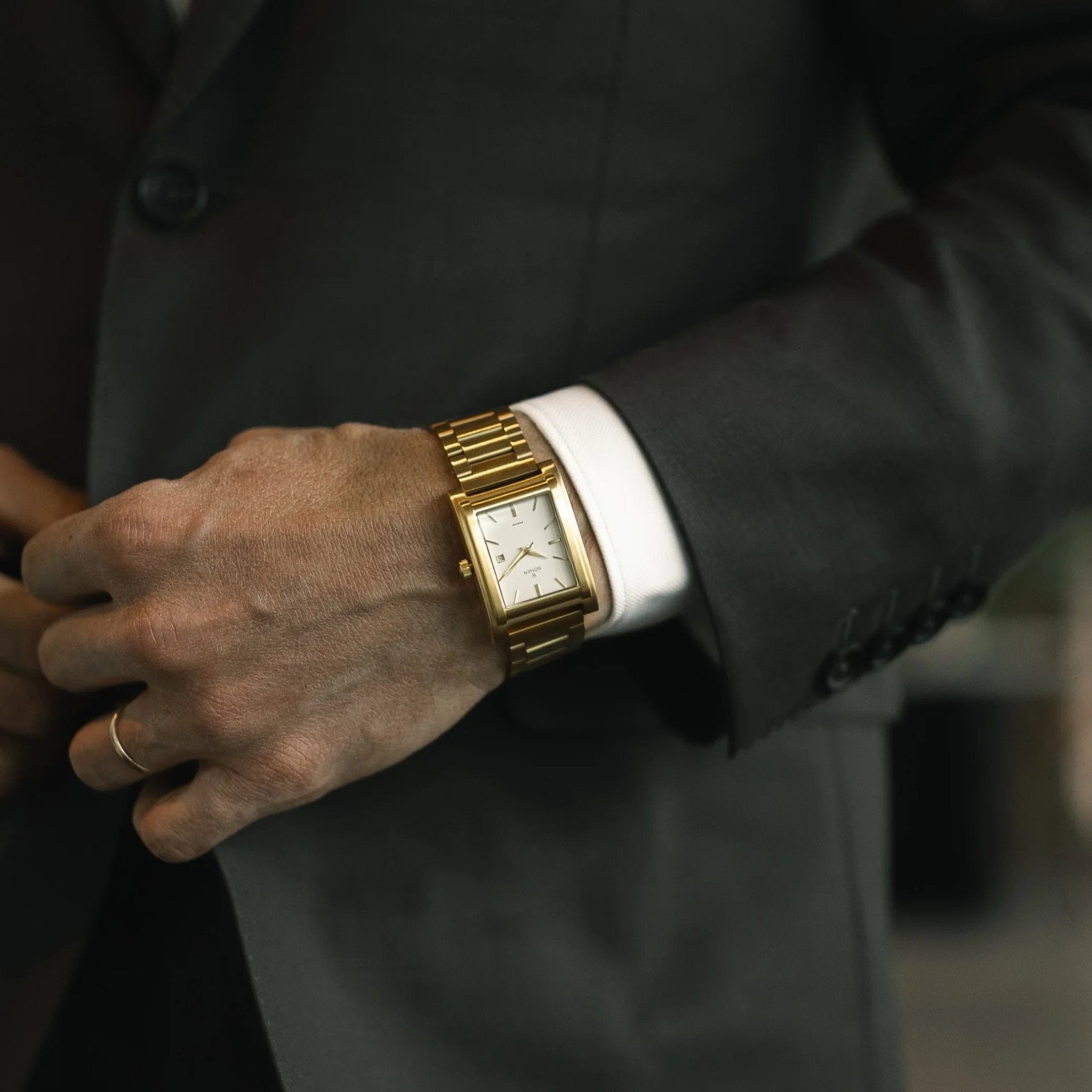 Gold-plated rectangular automatic watch with a white dial and gold bracelet, worn with a dark suit for an elegant, formal style.