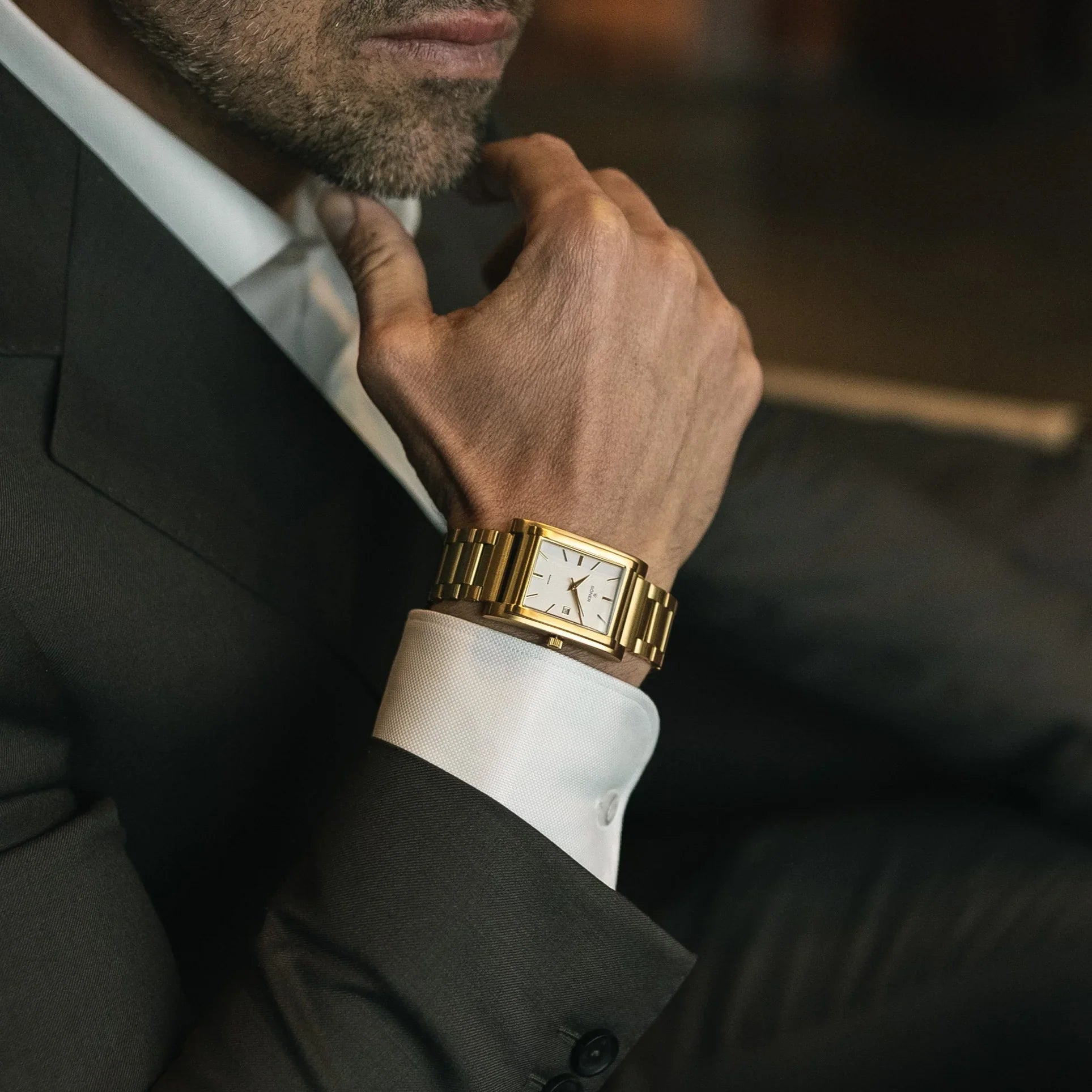Man wearing a gold-plated rectangular automatic watch with a white dial and gold bracelet, styled with a dark suit for a sophisticated, formal look.