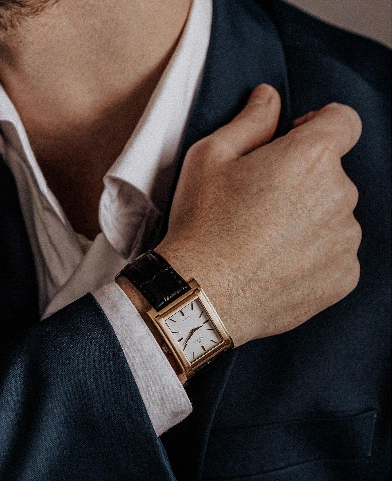 A person in a dark suit adjusts their white dress shirt cuff. They wear a watch with a rectangular gold case and black leather strap on their wrist. The background is softly blurred.