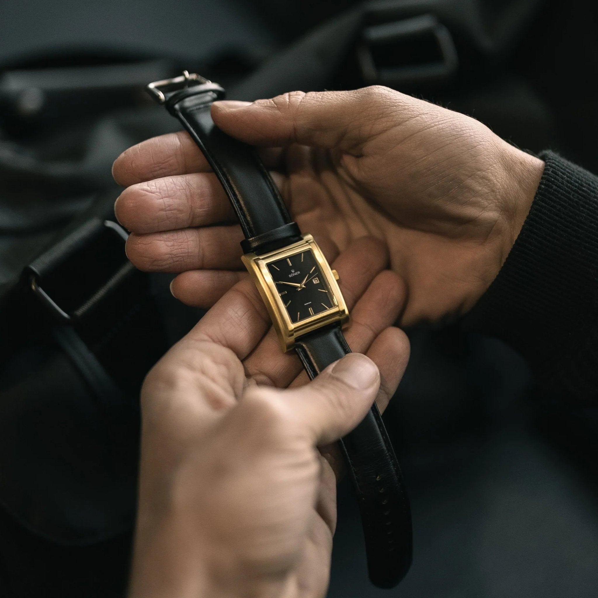 A brushed gold-plated Amorous Milano automatic rectangular watch with a black dial and leather strap, shown being held in hand for a close-up view of its elegant design.