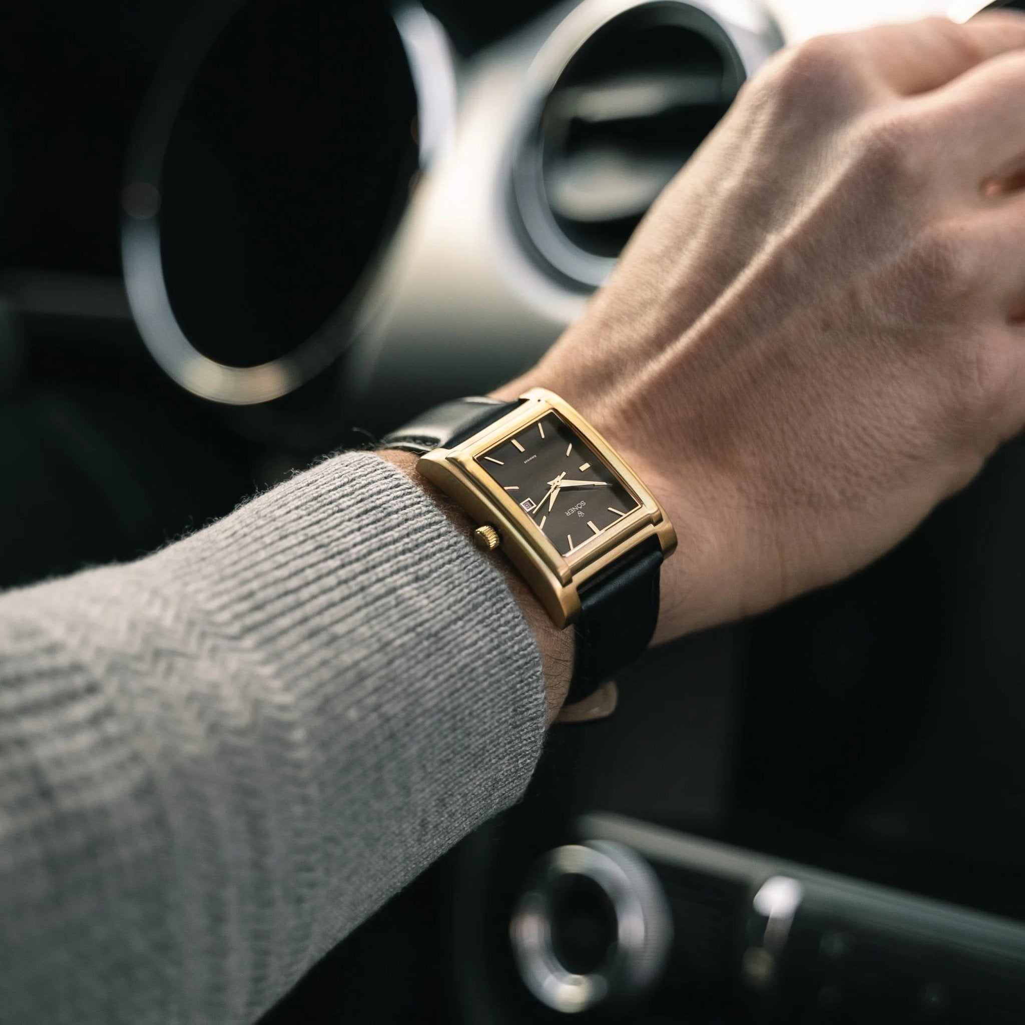 A brushed gold-plated rectangular Amorous Milano automatic watch with a black dial and leather strap, shown worn on the wrist while driving.