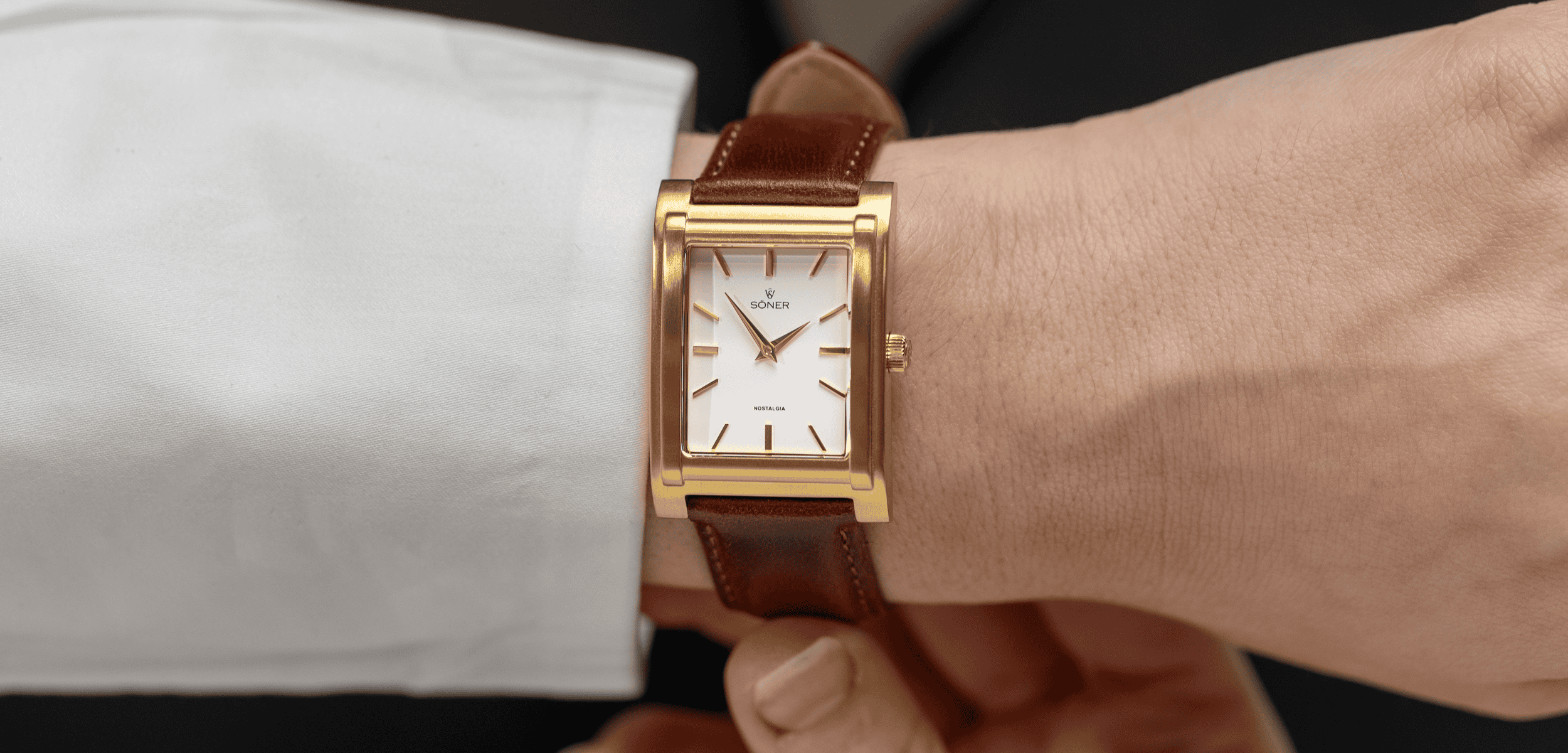 Close-up of a person wearing a stylish rectangular gold wristwatch with a white face and brown leather strap, showing the time as 10:10. The person is adjusting the watch strap with their other hand.