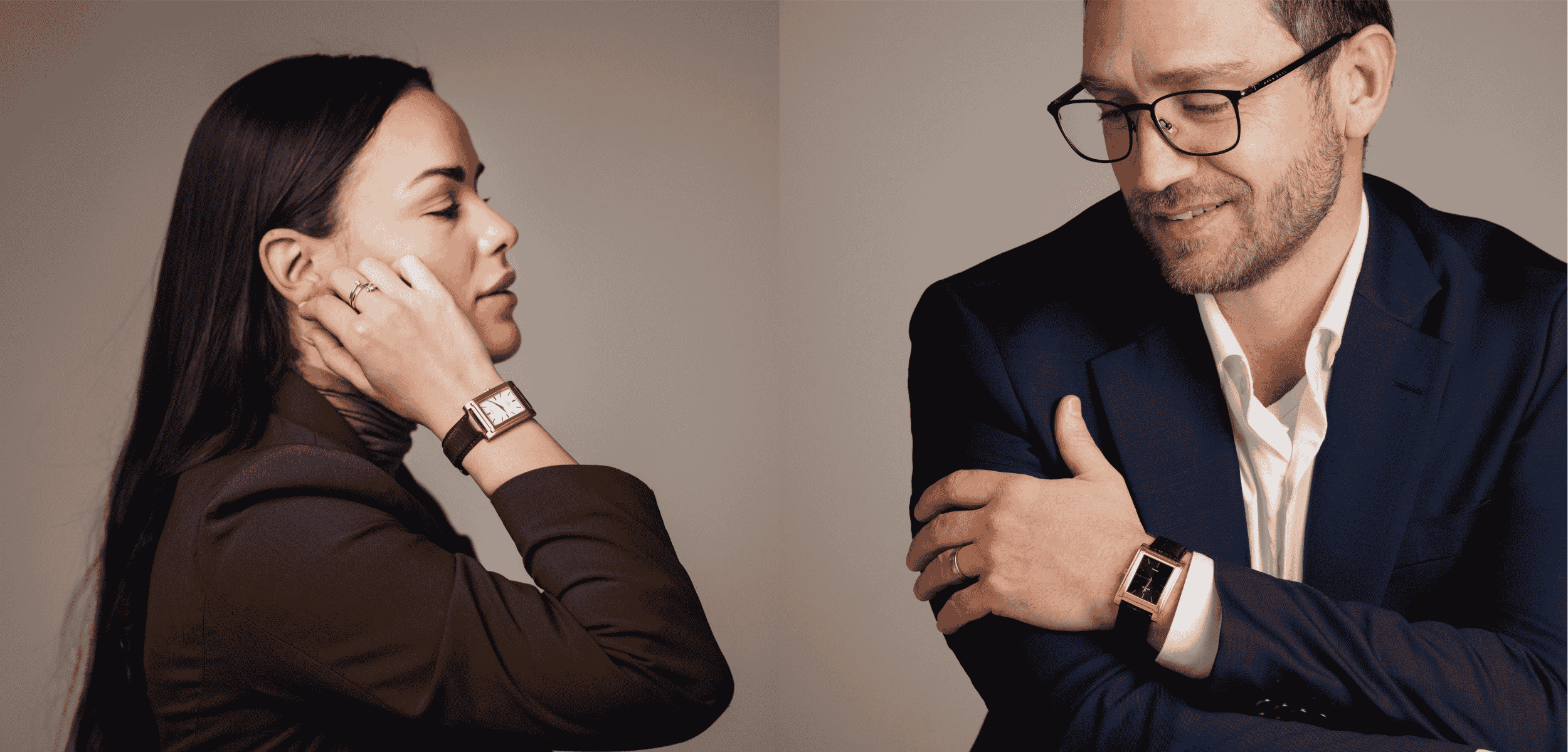 A woman and a man, both wearing stylish watches, are posed against a neutral background. The woman, wearing a brown blazer, touches her face, while the man, in a navy suit and glasses, crosses his arms and smiles.