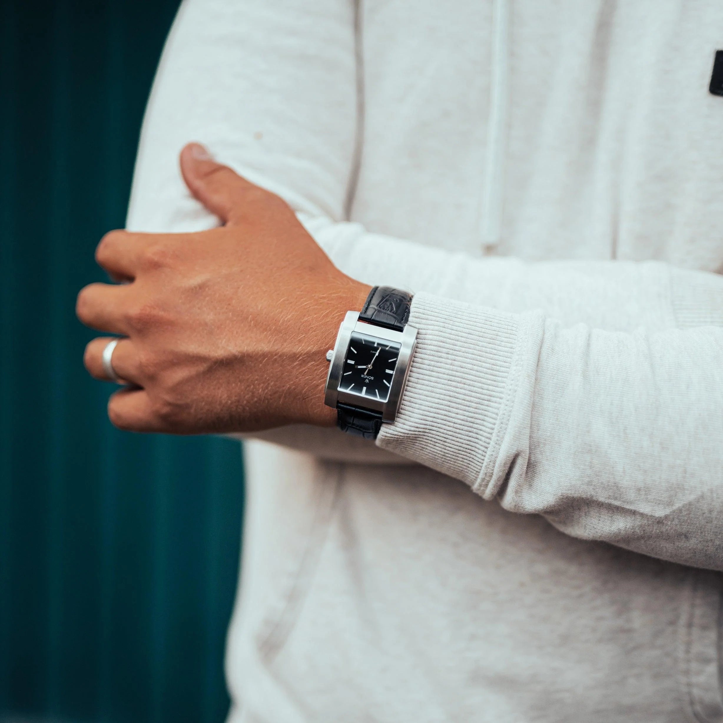 Close-up of a person wearing a light gray hoodie and a square black watch with a black leather strap on their wrist. The person has their arms crossed, showing a silver ring on one finger. The background is a blurred dark teal color.
