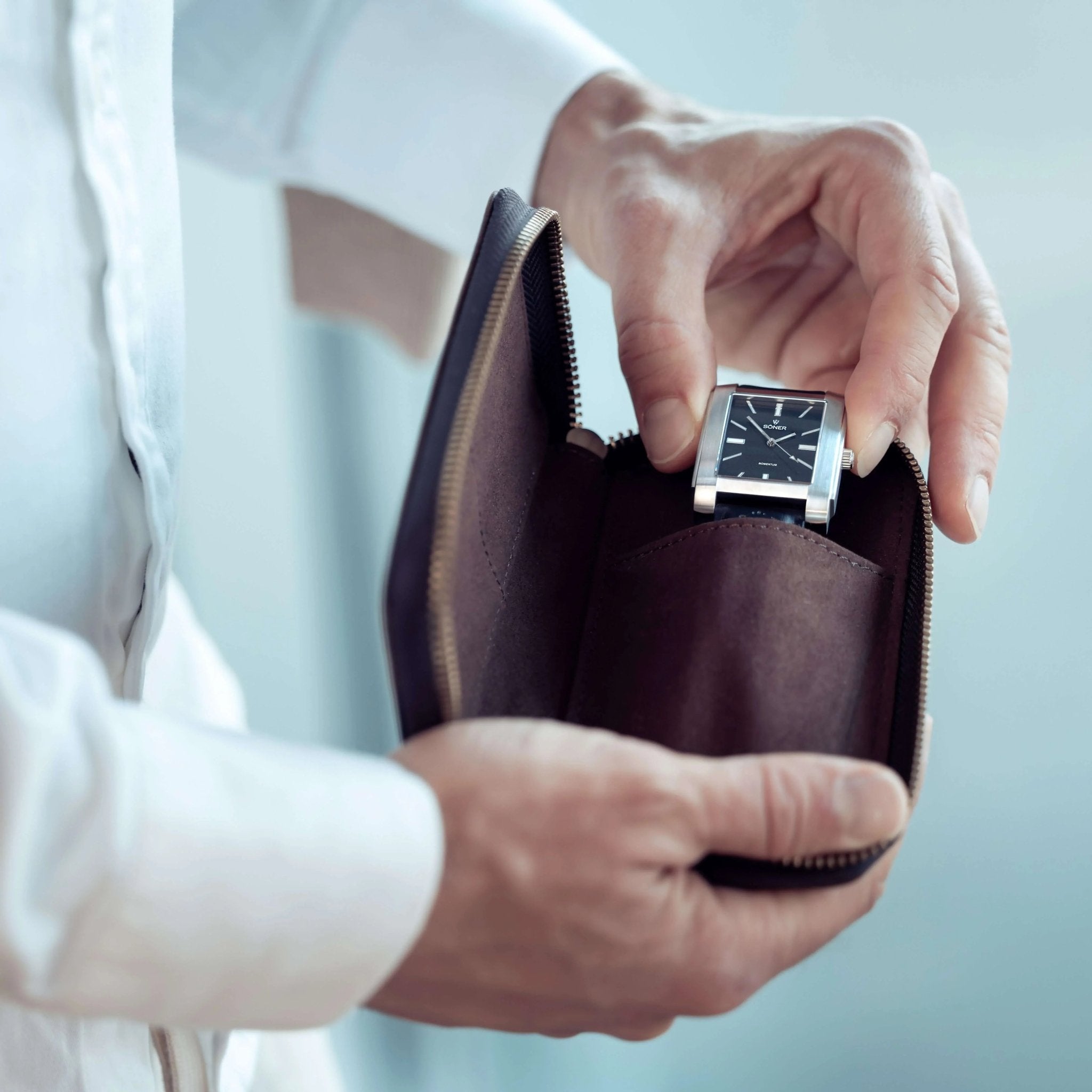 A person in a white shirt holds a Leather Watch Case, partially open to reveal a wristwatch inside. The watch features a square black face with silver accents.