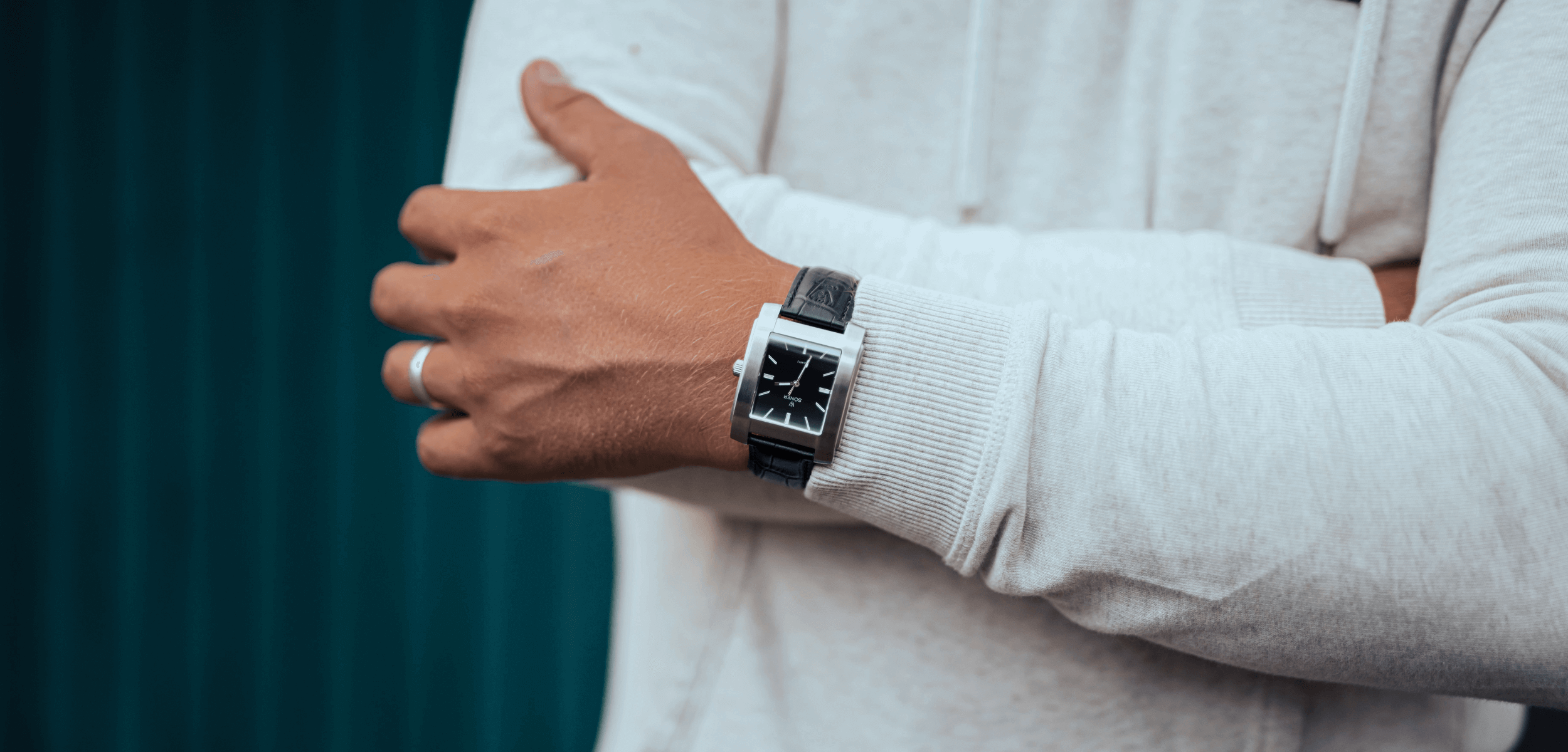 A person wearing a white hoodie crosses their arms, showcasing a black rectangular watch with silver edges. The timepiece features a geometric design and is secured by a sleek black band. Behind them, the backdrop is a dark teal corrugated surface.
