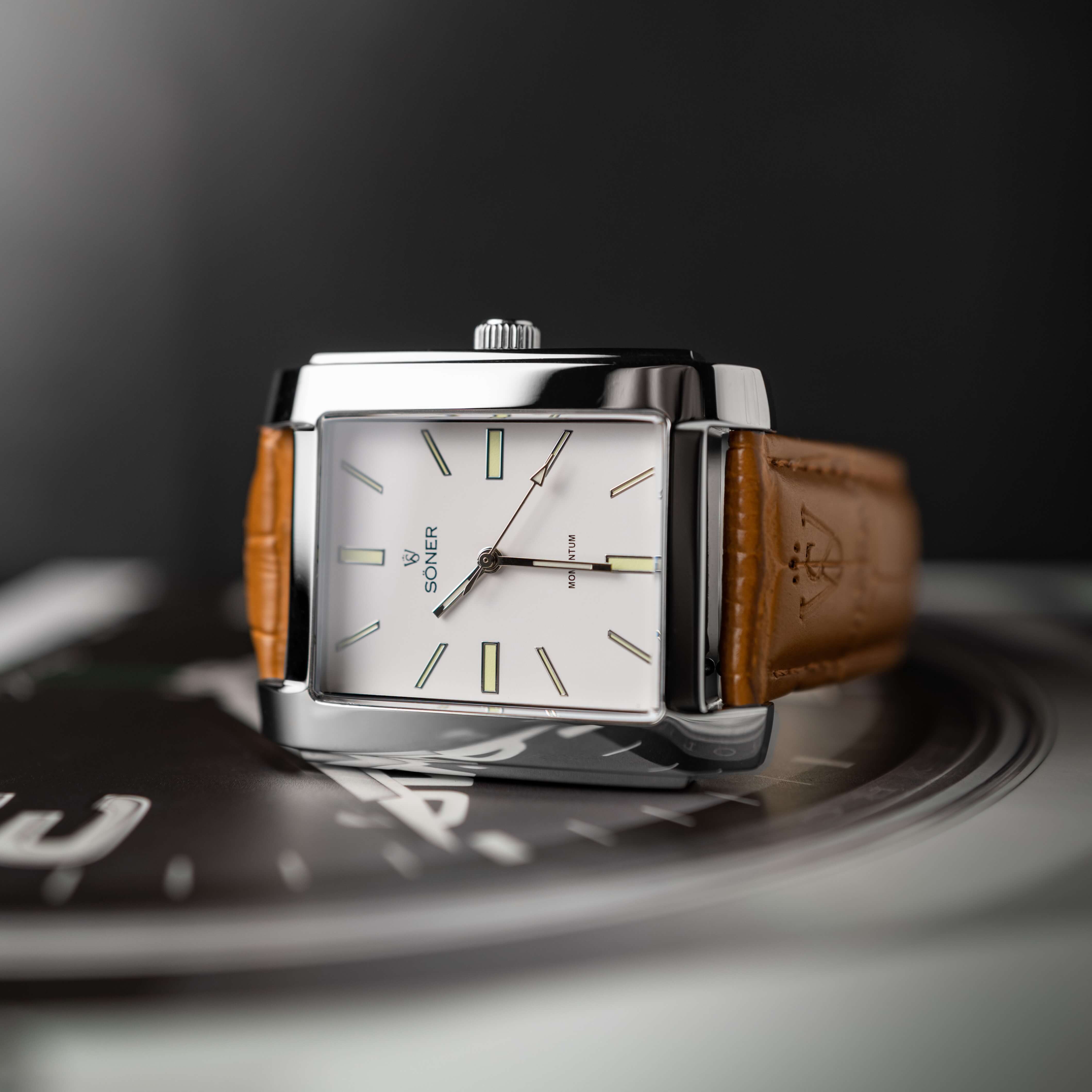 A stylish rectangular wristwatch with a white face, silver frame, and brown leather strap displayed on a glossy surface. It features minimalistic hour markers and simple hands, exuding an elegant and modern design.