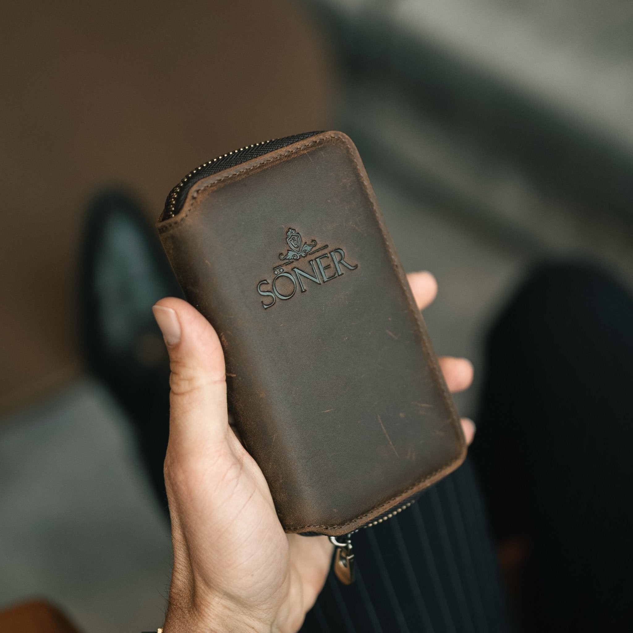 Hand holding a brown leather wallet with 'Soner' branding against a blurred background
