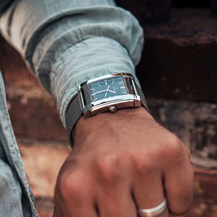A person in a light denim shirt holds a wrist adorned with a striking rectangular watch. The silver timepiece features a black face and metal mesh band, standing out against the softly blurred brick background.