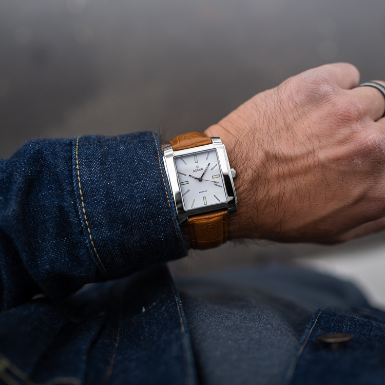 Close-up of a person wearing a rectangular watch with a silver case and brown leather strap. The timepiece features a white dial and silver hands. Draped in a denim jacket, the individual stands against a blurred background.