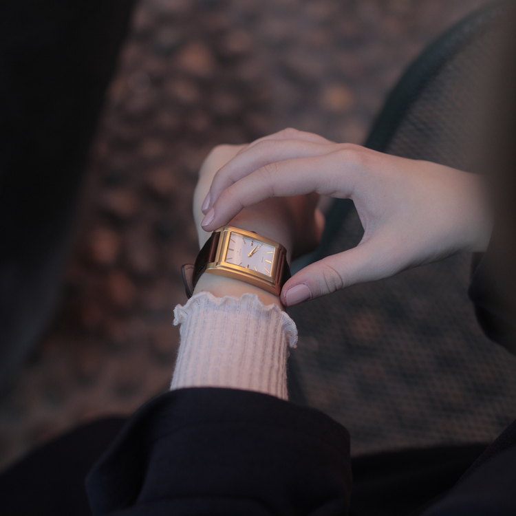 A person with a rectangular watch featuring a gold face and black band checks the time. They wear a dark coat over a light-colored, ribbed sleeve. The background is softly blurred.