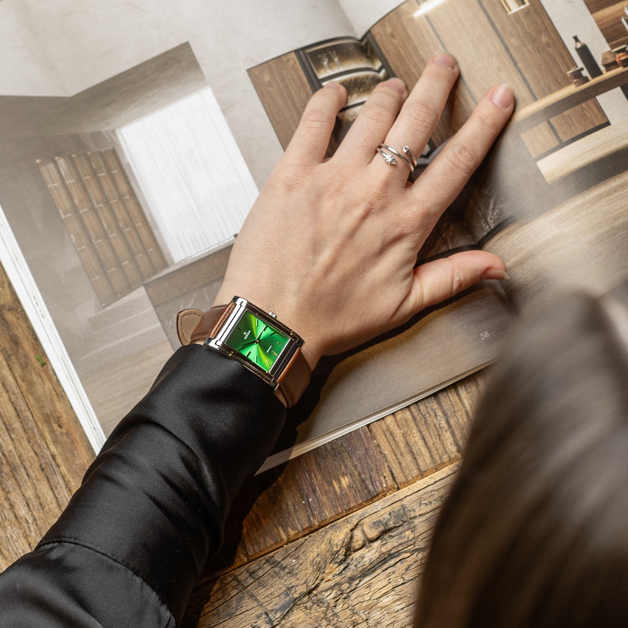 A person with a green rectangular smartwatch on their wrist turns the page of a furniture magazine. The individual is wearing a black satin long-sleeve shirt and a silver knotted ring, all set against the warm texture of a wooden table.