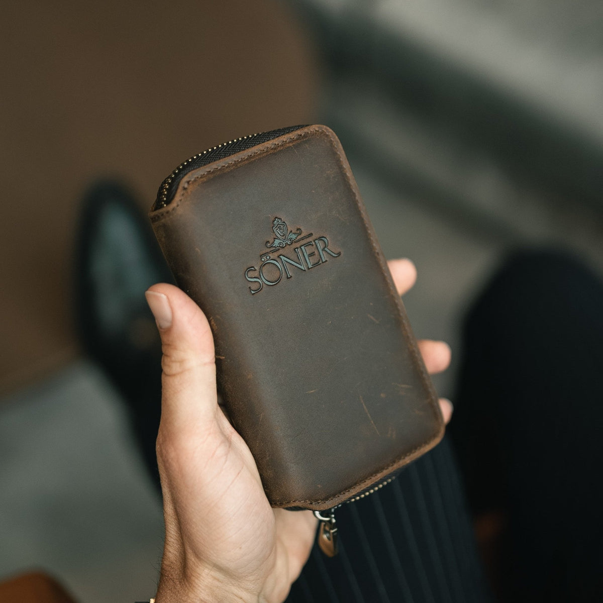 A hand holding a brown leather watch travel case with a zipper, embossed with the brand name SONER and a small decorative emblem above the text.