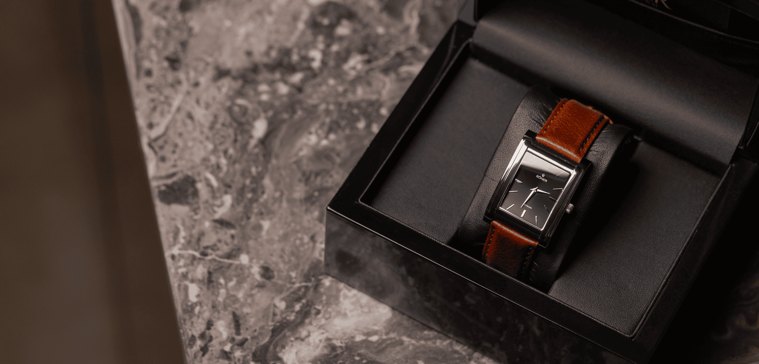 A rectangular wristwatch with a brown leather strap sits in a black presentation box on a marbled surface. The watch face is black with white lines indicating the hours, and silver minute and hour hands.