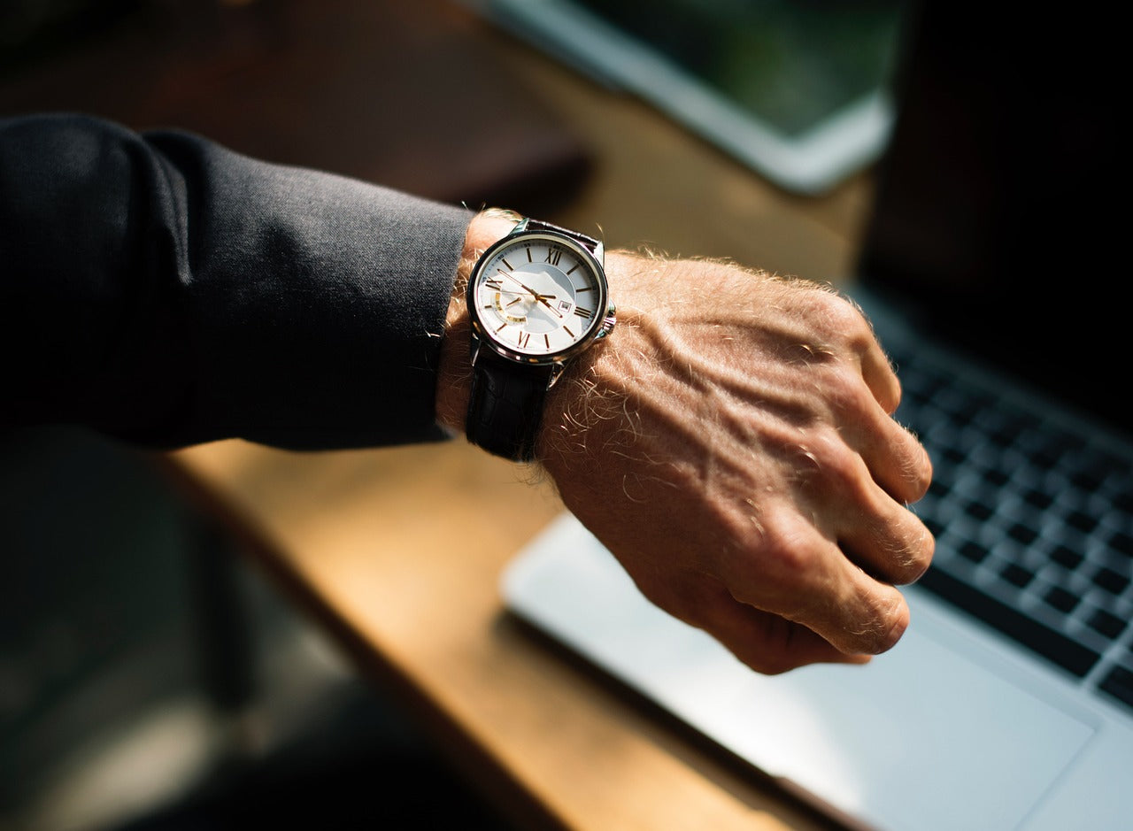 A person in a dark suit adjusts their jacket, wearing a gold wristwatch and a wedding ring on their left hand. The background is softly blurred.