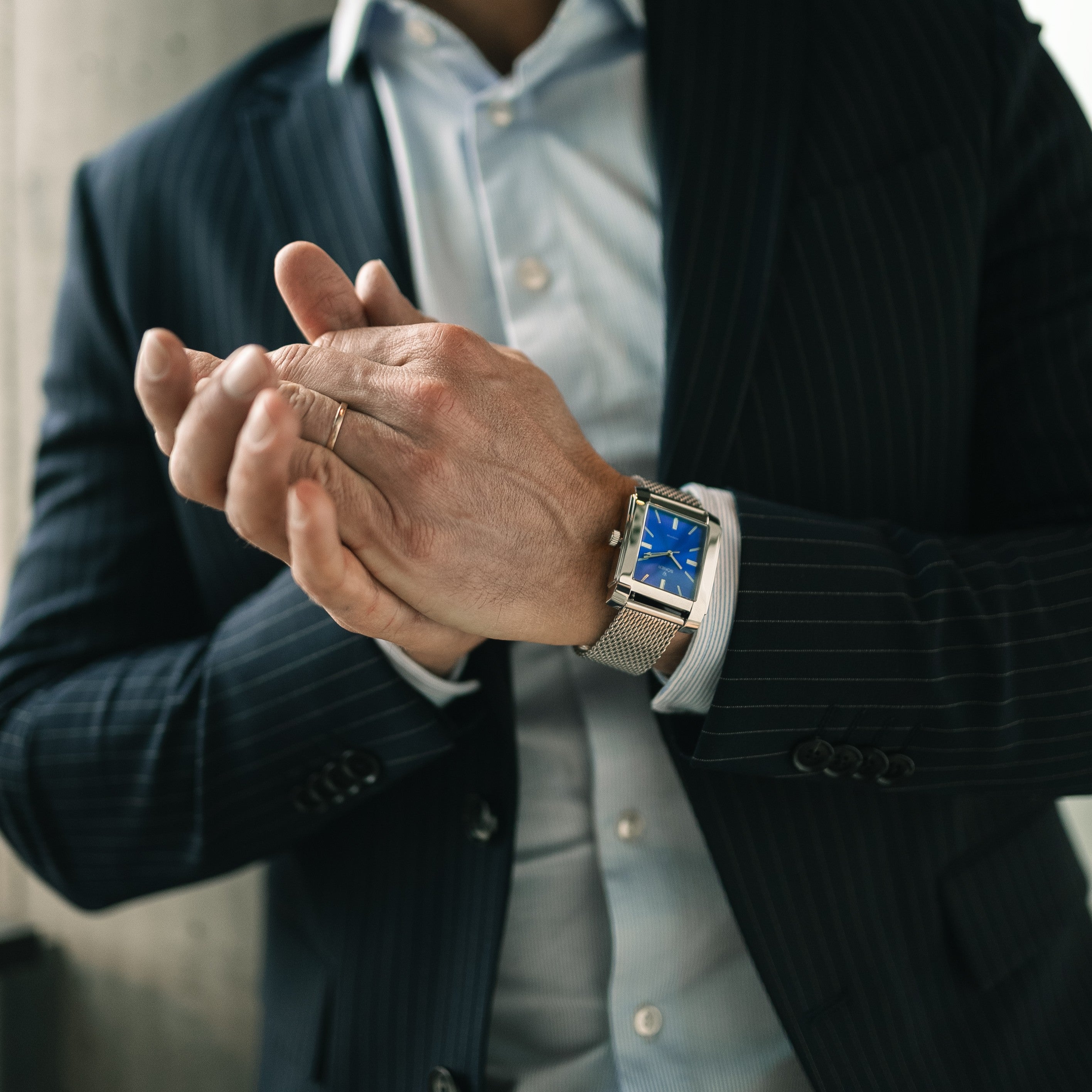 A person in a dark suit adjusts their jacket, wearing a gold wristwatch and a wedding ring on their left hand. The background is softly blurred.