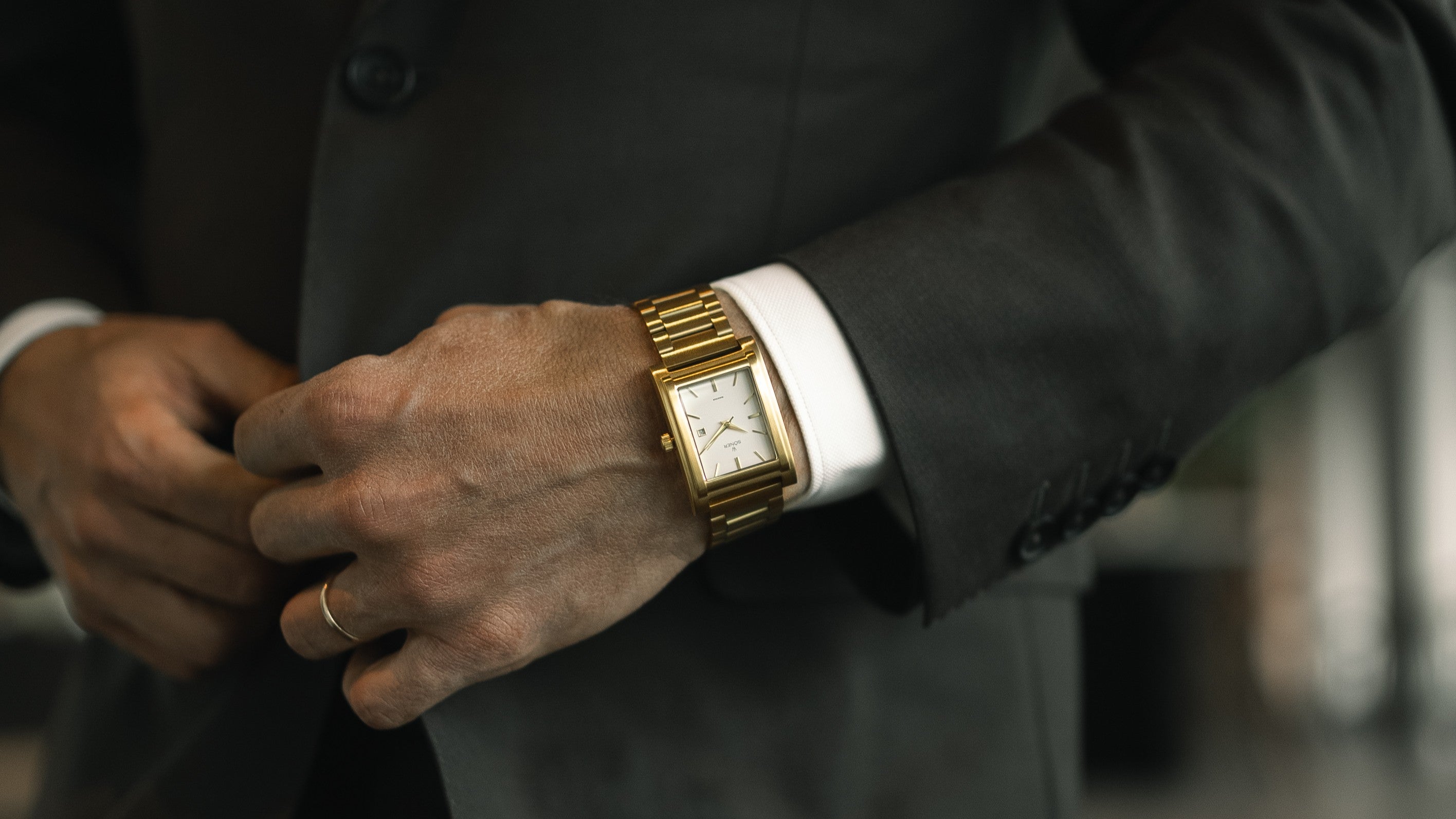 A close-up of a person in a dark suit adjusting their jacket, wearing a gold wristwatch and a wedding ring on their left hand.
