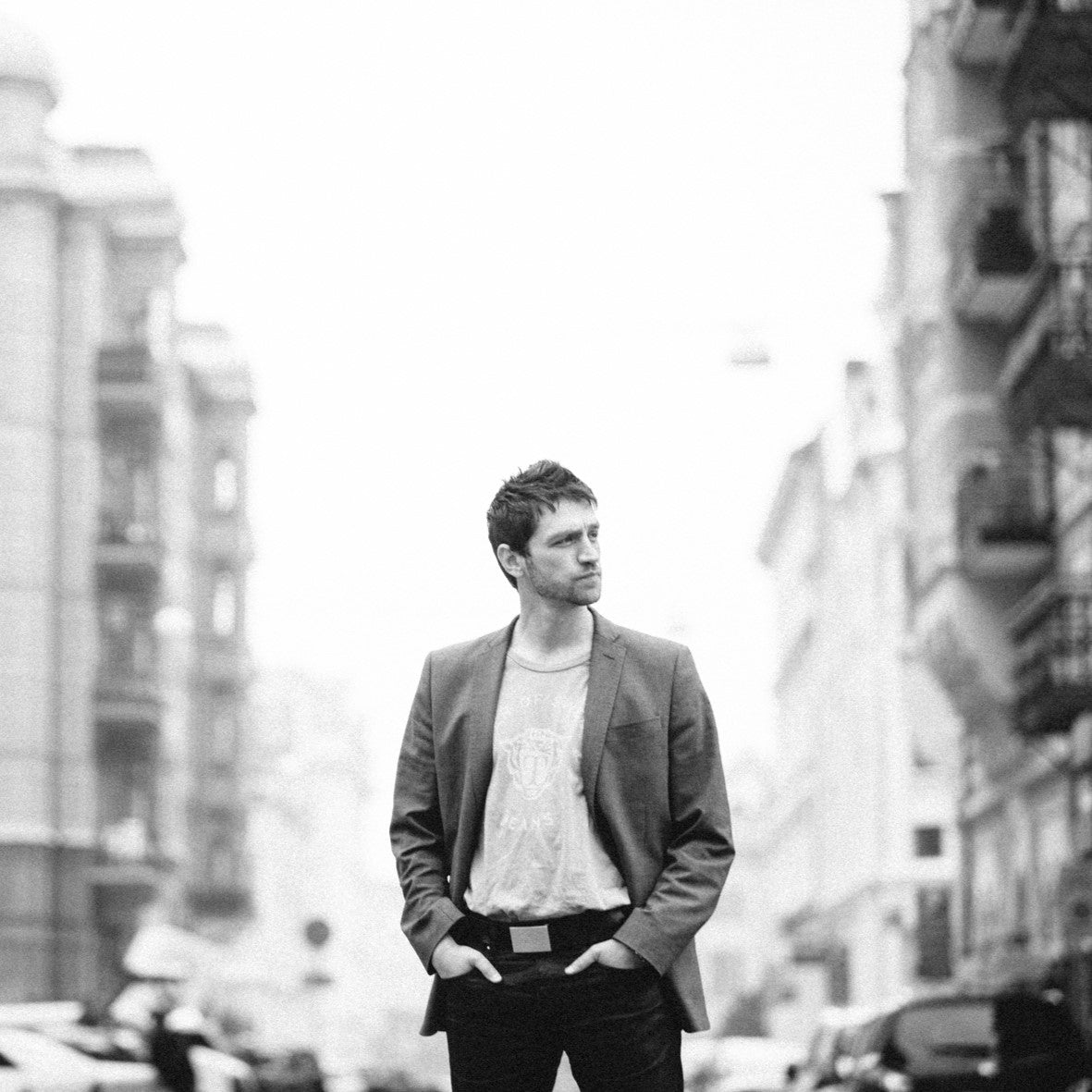 A man stands confidently on a city street, hands in his pockets. Hes wearing a casual t-shirt with a blazer over it, and a sleek rectangular watch glints subtly on his wrist. The street is slightly blurred, suggesting a bustling urban environment. The image is in black and white.