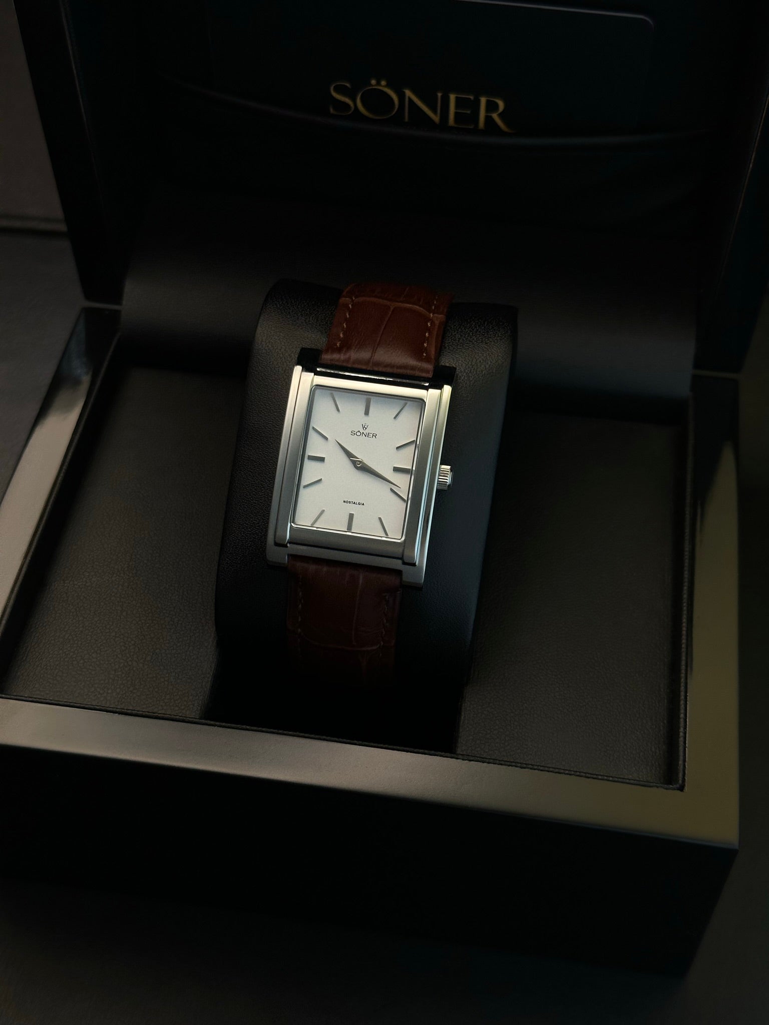 A luxury wristwatch with a brown leather band and a rectangular silver case is displayed in a black Soner box. The watch has a white face with minimalistic hour markers and sleek hands.