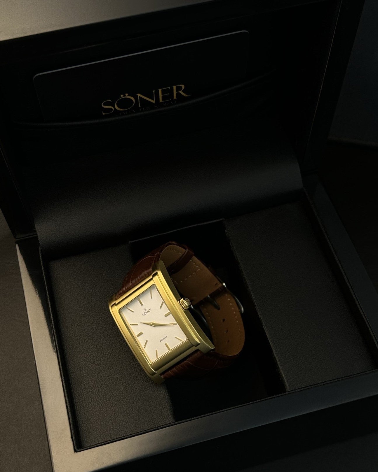 A rectangular gold watch with a white face and brown leather strap is elegantly showcased in an open black box, where the brand name SÖNER graces the inside lid. This exquisite timepiece, a testament to minimalistic design, features subtle markings that enhance its refined allure.