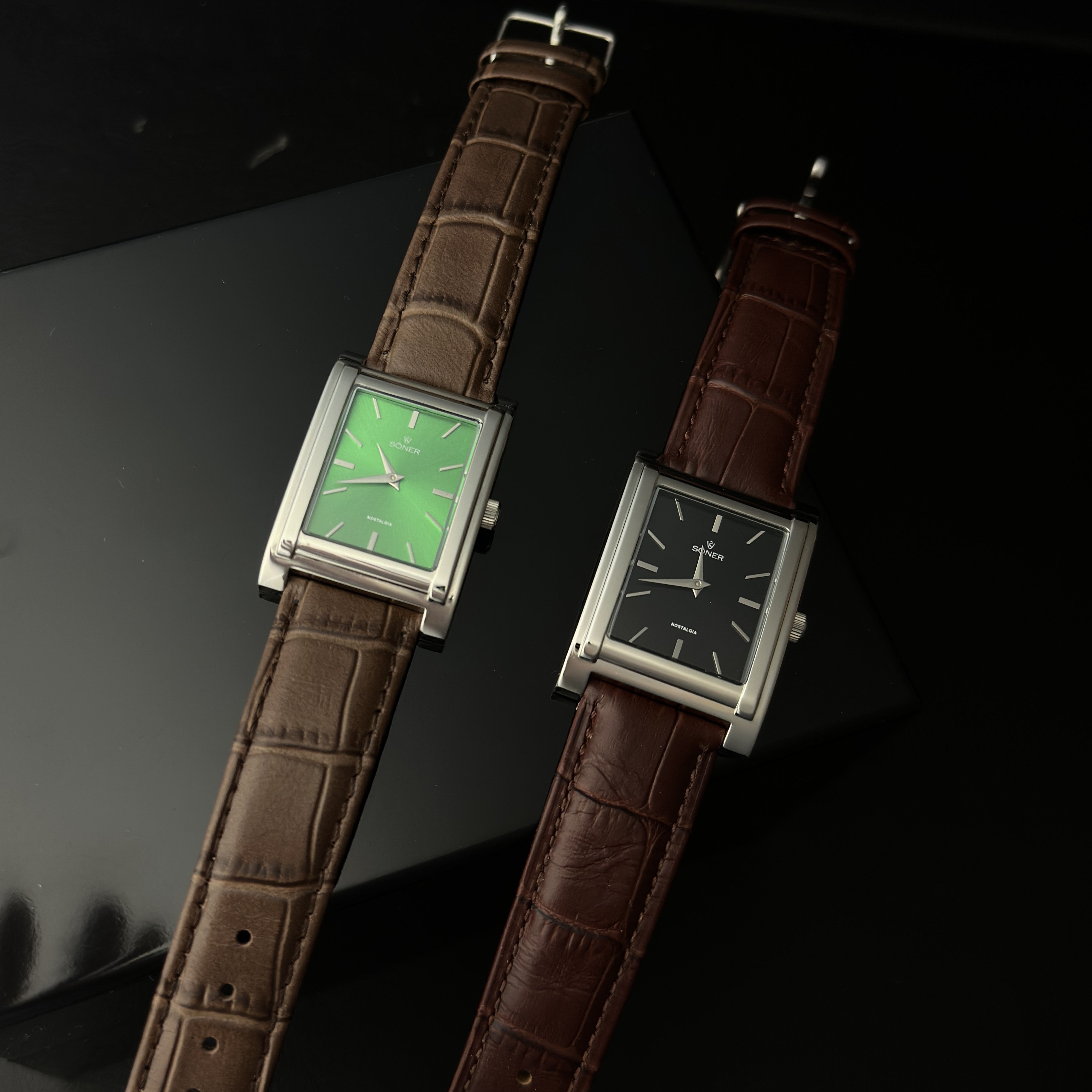 Two rectangular wristwatches with leather straps are displayed side by side on a dark surface. The left watch has a green face and brown strap, while the right watch features a black face and brown strap. Both have minimalist designs.